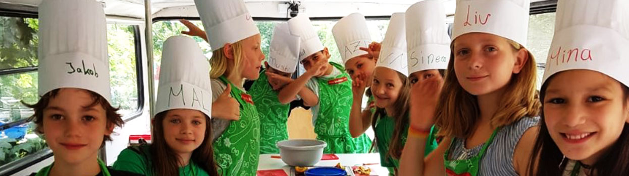 10 Schüler stehen im Rosa-Parks-Bus. Sie gucken und lächeln in die Kamera. Die Kinder tragen grüne Kochschürzen und weiße Kochmützen mit ihren Namen drauf.