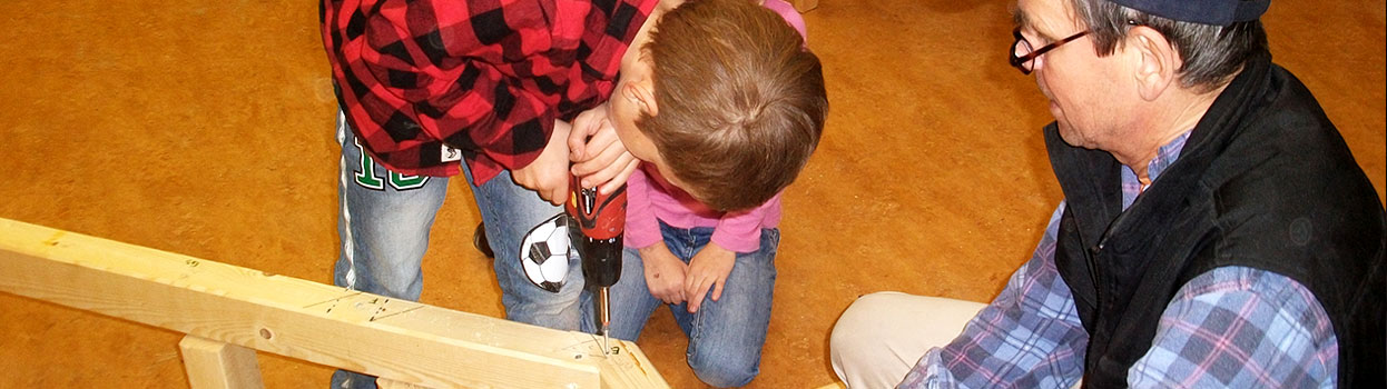 Ein Schulkind schraubt mit einem Akkuschrauber eine Schraube in eine Holzverbindung. Daneben sitzt ein Lehrer, passt auf und unterrichtet den Jungen.