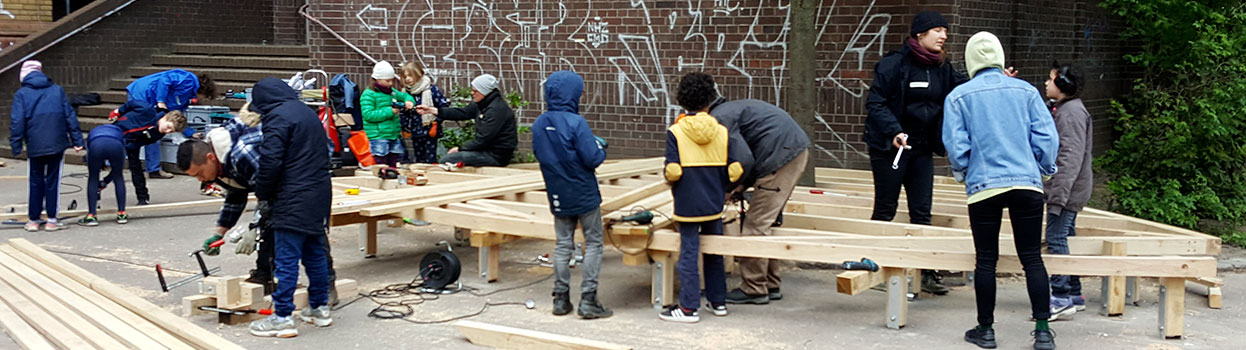 Schüler haben auf dem Schulhof unterricht. Die Schüler arbeiten an einem Holzpodest. Einige Schüler schrauben, andere Schüler sägen und andere Kinder unterstützen.