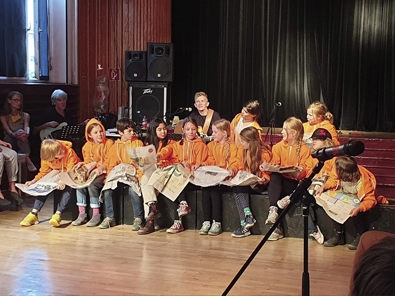 12 Kinder spielen ein Theaterspiel. Sie sitzen in einer Reihe. Alle halten Zeitungen in den Händen. Sie gucken zu einer Schülerin. Die eine Schülerin sitzt in der Mitte. Eine Lehrerin sitzt hinter den Kindern. Die Lehrerin spielt dabei Gitarre.