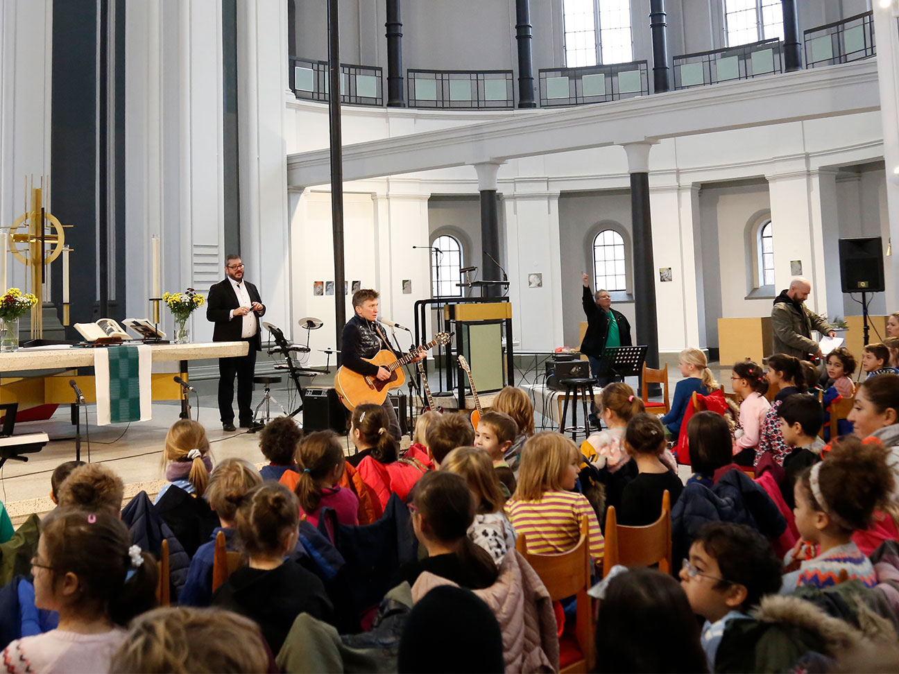 In einer Kirche findet ein Schulkonzert statt. Vorne steht eine Lehrerin. Die Lehrerin singt und spielt Gitarre. Die Kirche ist voll mit Kindern. Alle Kinder hören der Lehrerin zu.