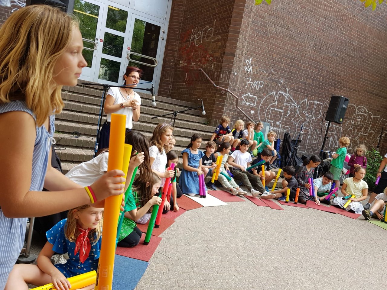Auf dem Schulhof wird Musik gemacht. Die Schulkinder spielen Klangrohre. Sie sitzen in einem Kreis.