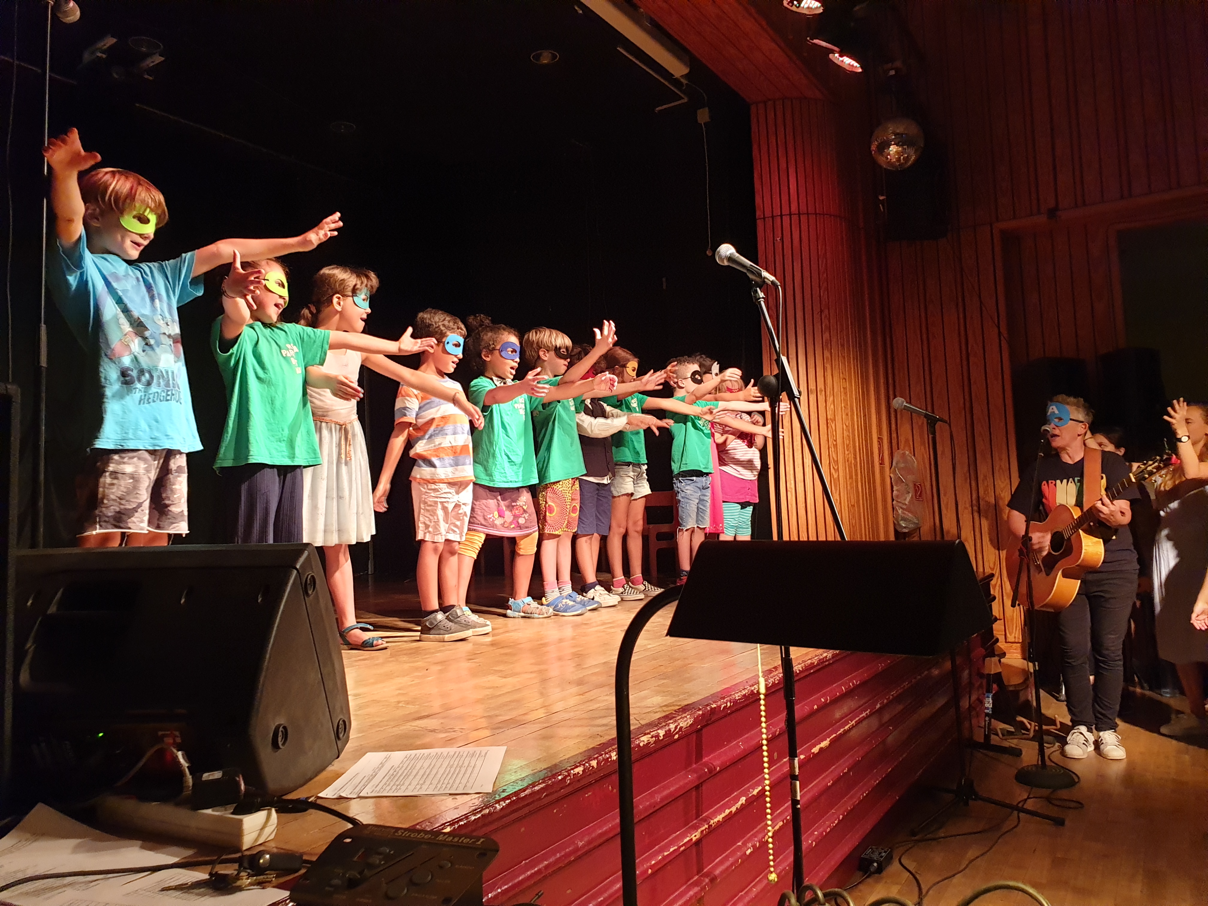 11 junge Schulkinder stehen auf einer Bühne. Sie sind bunt gekleidet und tragen bunte Augenmasken. Die Kinder singen laut und heben ihre Hände in die Luft. An der Seite steht eine Lehrerin. Die Lehrerin singt mit und spielt Gitarre.