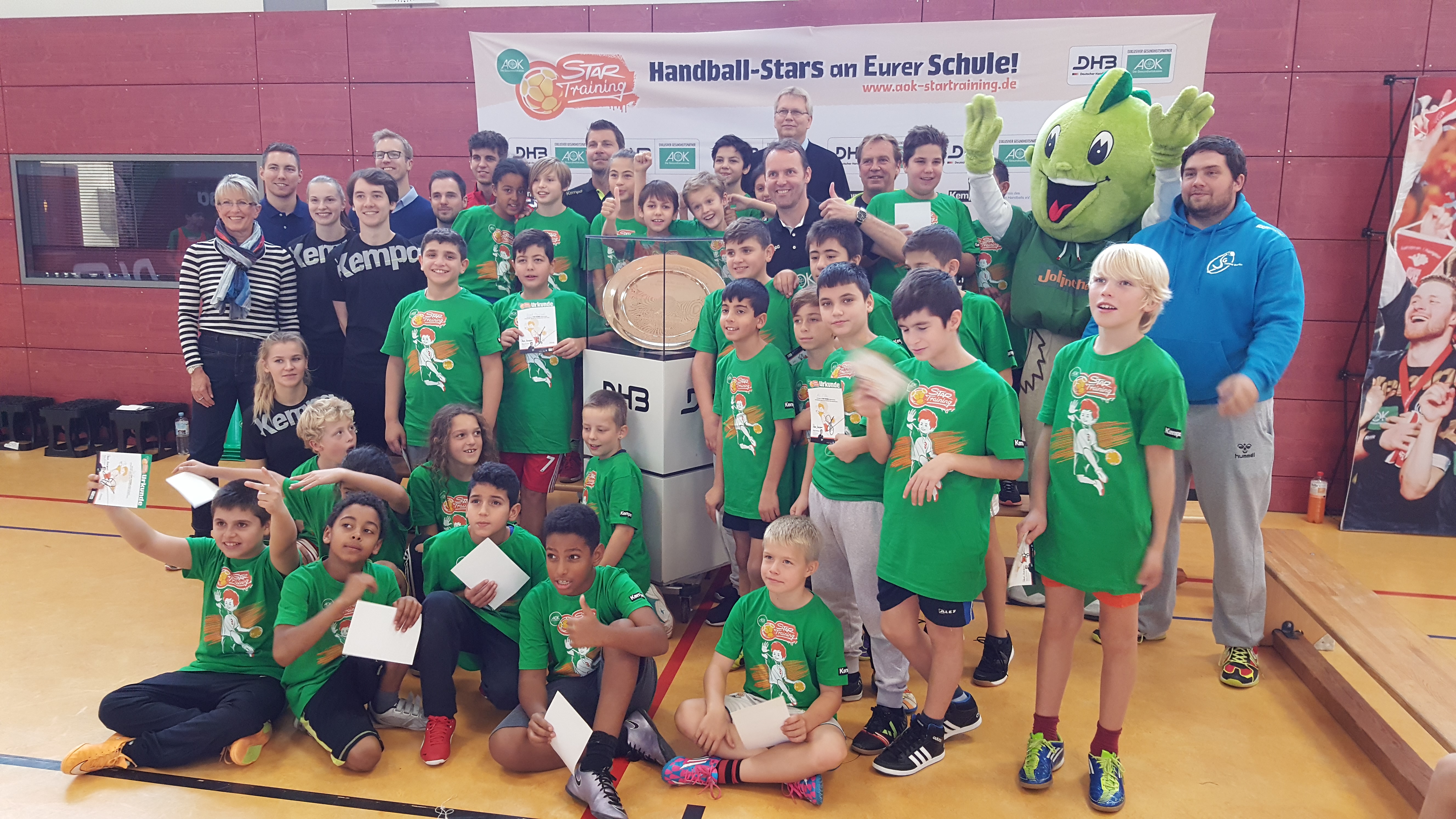 Die glücklichen Handball-Stars unserer Schule. Unser Handball-Team steht vor einem Pokal. Der Pokal glänzt und hat die Form eines großen Tellers. Die Kinder halten Urkunden hoch. Hinten stehen Lehrer und ein grünes Maskottchen.
