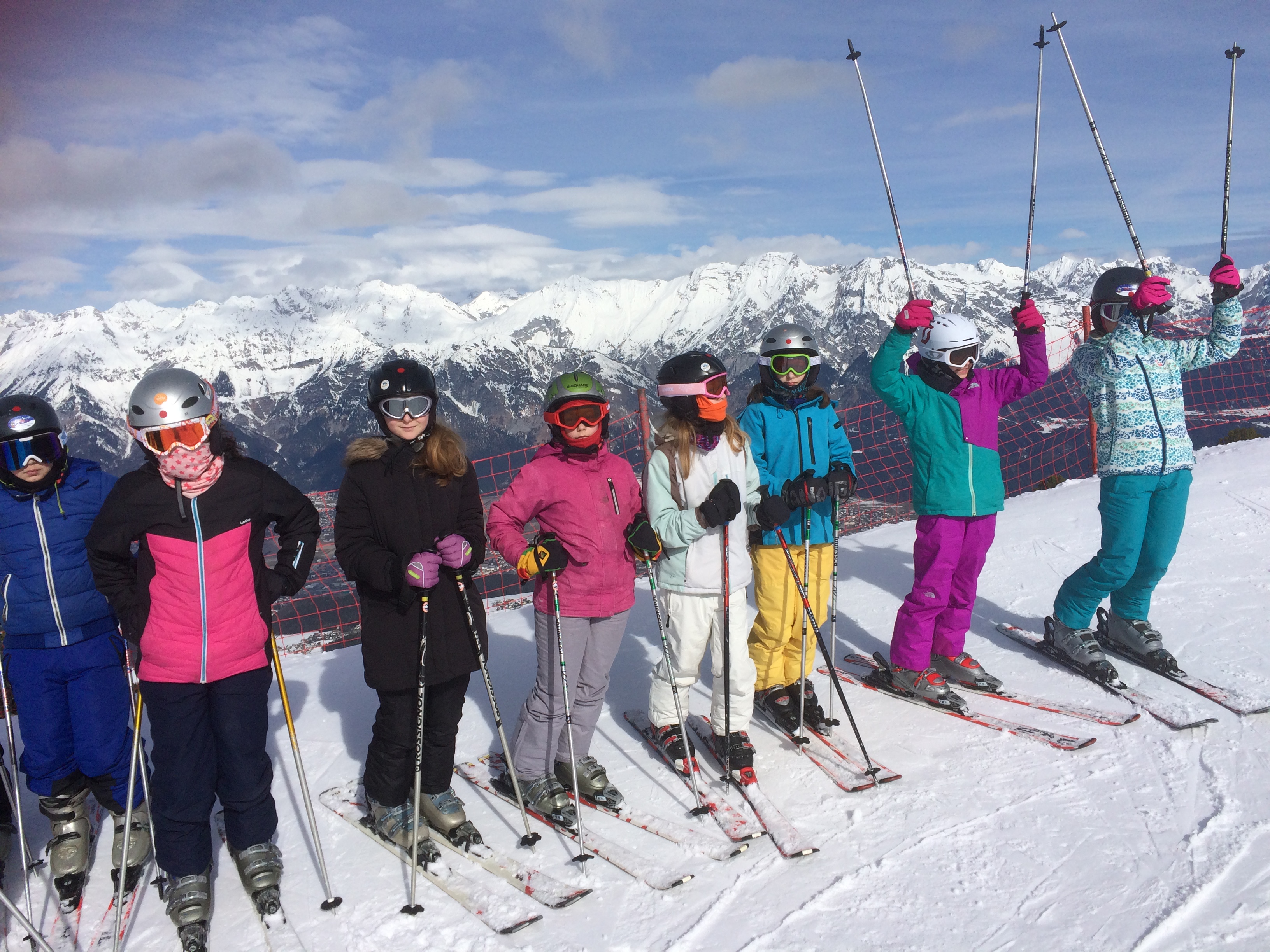 Auf einer Klassenfahrt. Acht Schüler stehen mit Ski-Anzügen, Skiern, Helmen und Schutzbrillen nebeneinander. Hinter den Kindern sind riesige Gebirge.