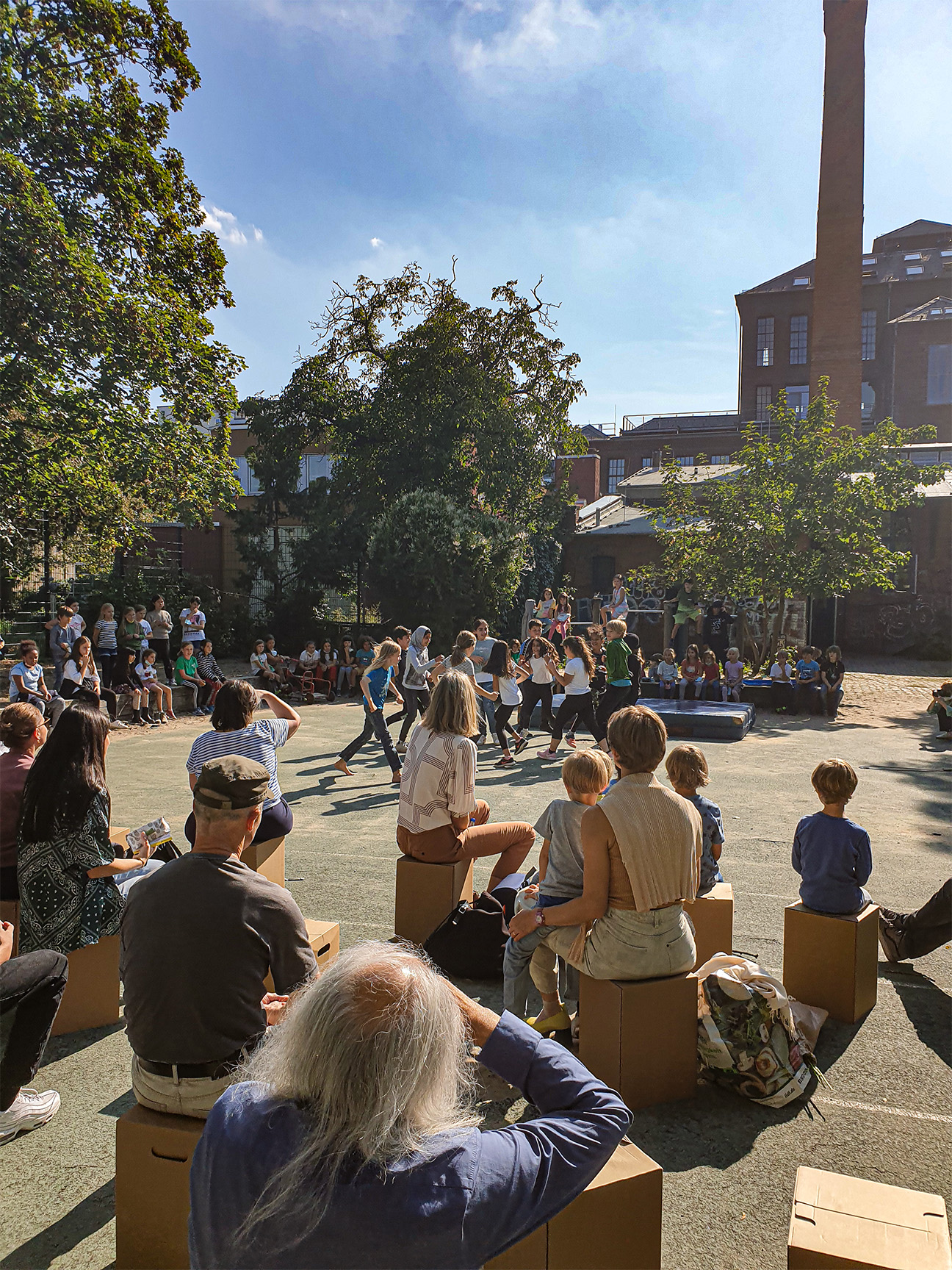 Eine Aufführung auf dem Schulhof. Kinder tanzen in einer Mitte. Herum sitzen mehr Kinder und gucken zu.