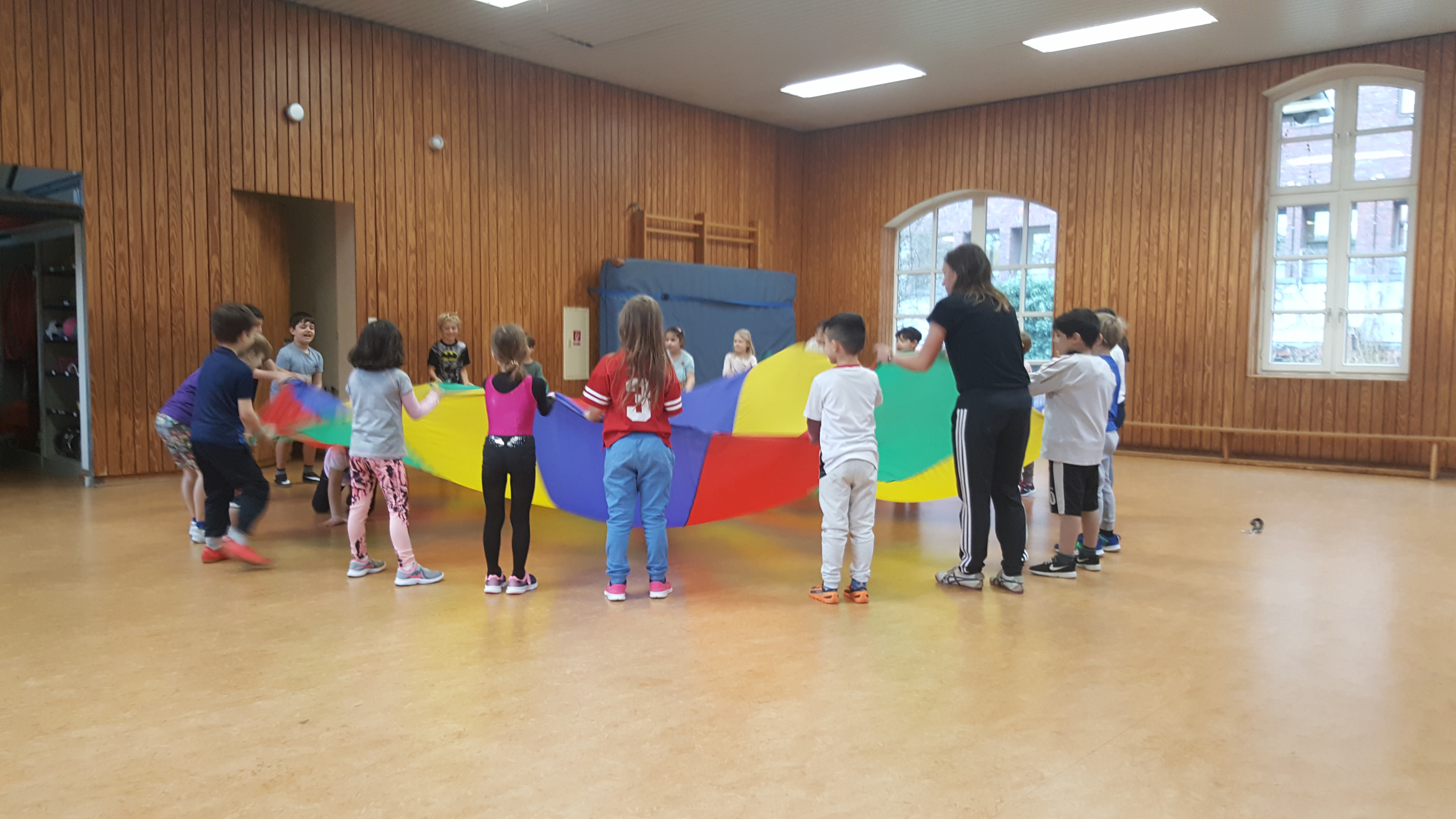 Unterricht in der Sporthalle. Viele Kinder halten und schwingen ein buntes Schwingtuch. Eine Lehrerin hilft dabei. Unter dem Schwingtuch krabbelt eine Schülerin von der einen Seite, zur anderen Seite.