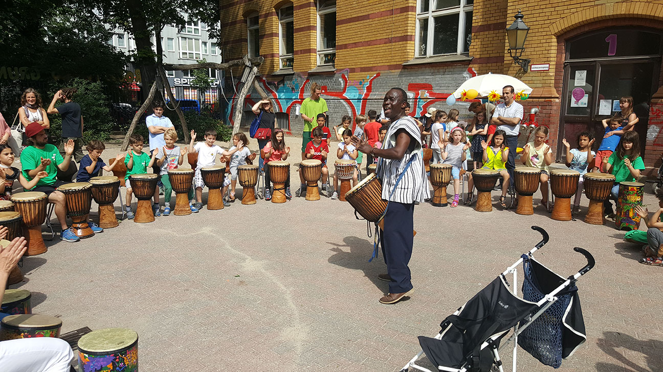 Auf dem Schulhof trommeln Kinder im Kreis, während sie von einem Trommel-Lehrer begleitet werden.