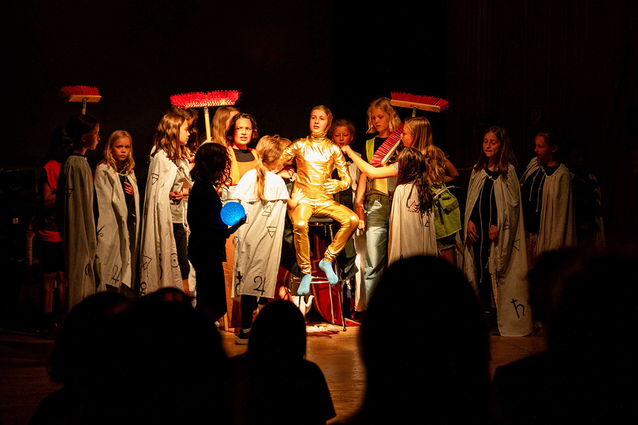 Ein Schulauftritt gespielt von Schulkindern. In der Mitte sitzt ein Schüler im goldenen Kostüm, rundherum stehen viele Schüler in hellen Umhängen. Ein paar Schüler halten besen.