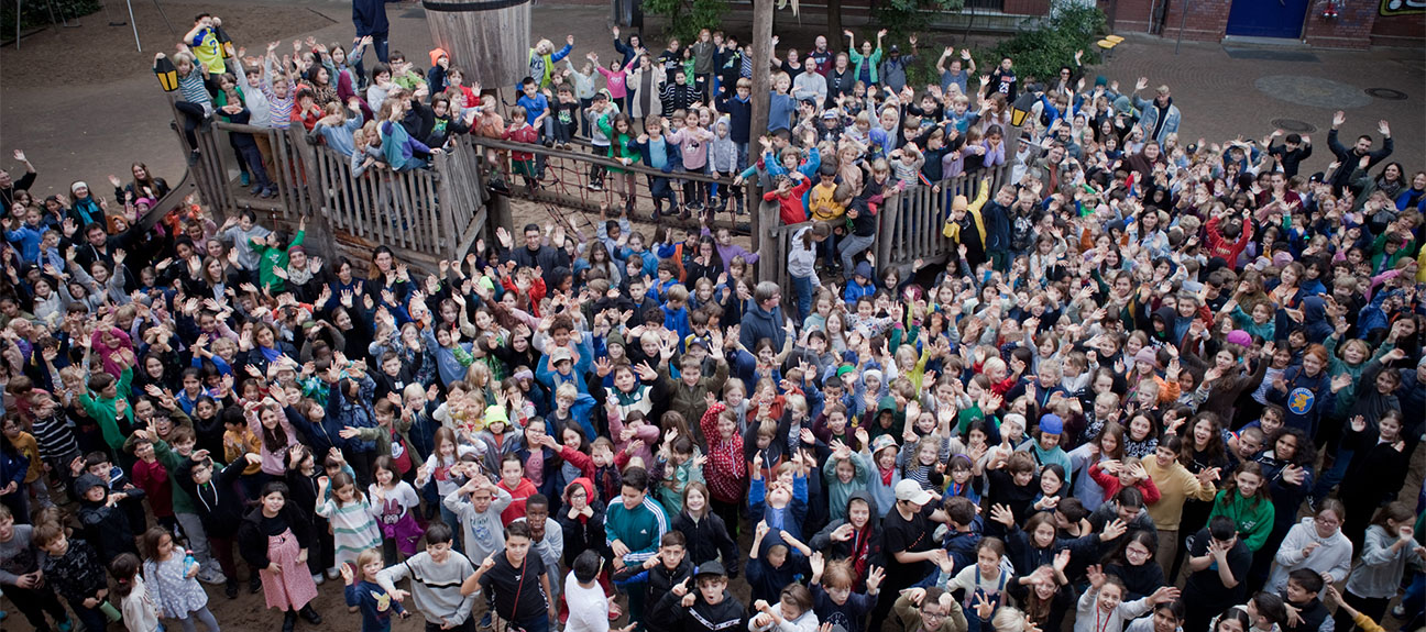 Eine Drohnenaufnahme über unserem Schulhof. Viele versammelte Kinder und Lehrer gucken nach oben und winken der Drohne zu.